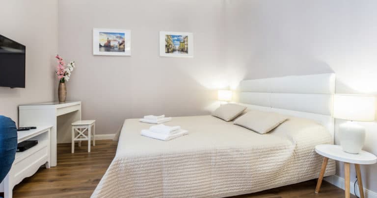 Bright bedroom with double bed, beige quilt, folded towels, wall-mounted TV, and small desk.