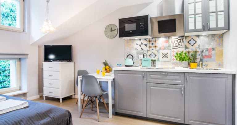 Studio kitchen area with grey cabinets, small dining table, TV and adjacent bed.