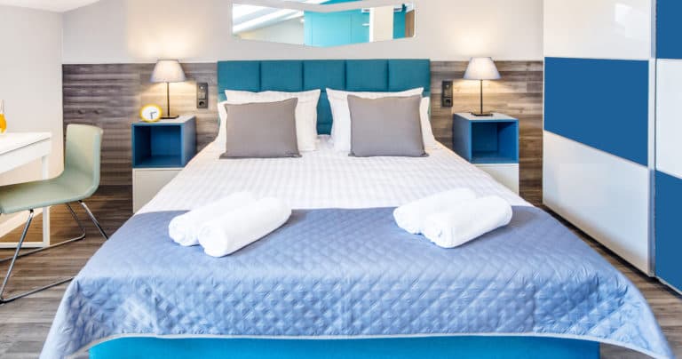 Front-facing double bed with blue quilt, three rolled towels, bedside tables, and soft lighting.