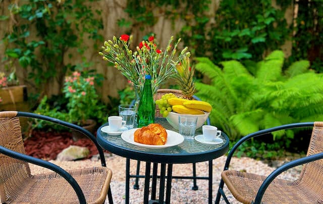 Bistro table in garden set with croissant, coffee cups, fruit bowl and tall flower vase